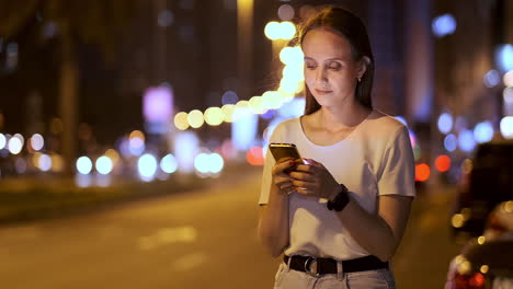 Una-Hermosa-Joven-En-Verano-En-Una-Gran-Ciudad-Nocturna-Mantiene-Un-Teléfono-Inteligente-En-Su-Interior-Y-Escribe-Un-Mensaje-Lee-Correos-Electrónicos-Y-Se-Comunica-En-Salas-De-Chat-Y-Redes-Sociales-Mantiene-Un-Blog-Mira-La-Pantalla-Del-Teléfono